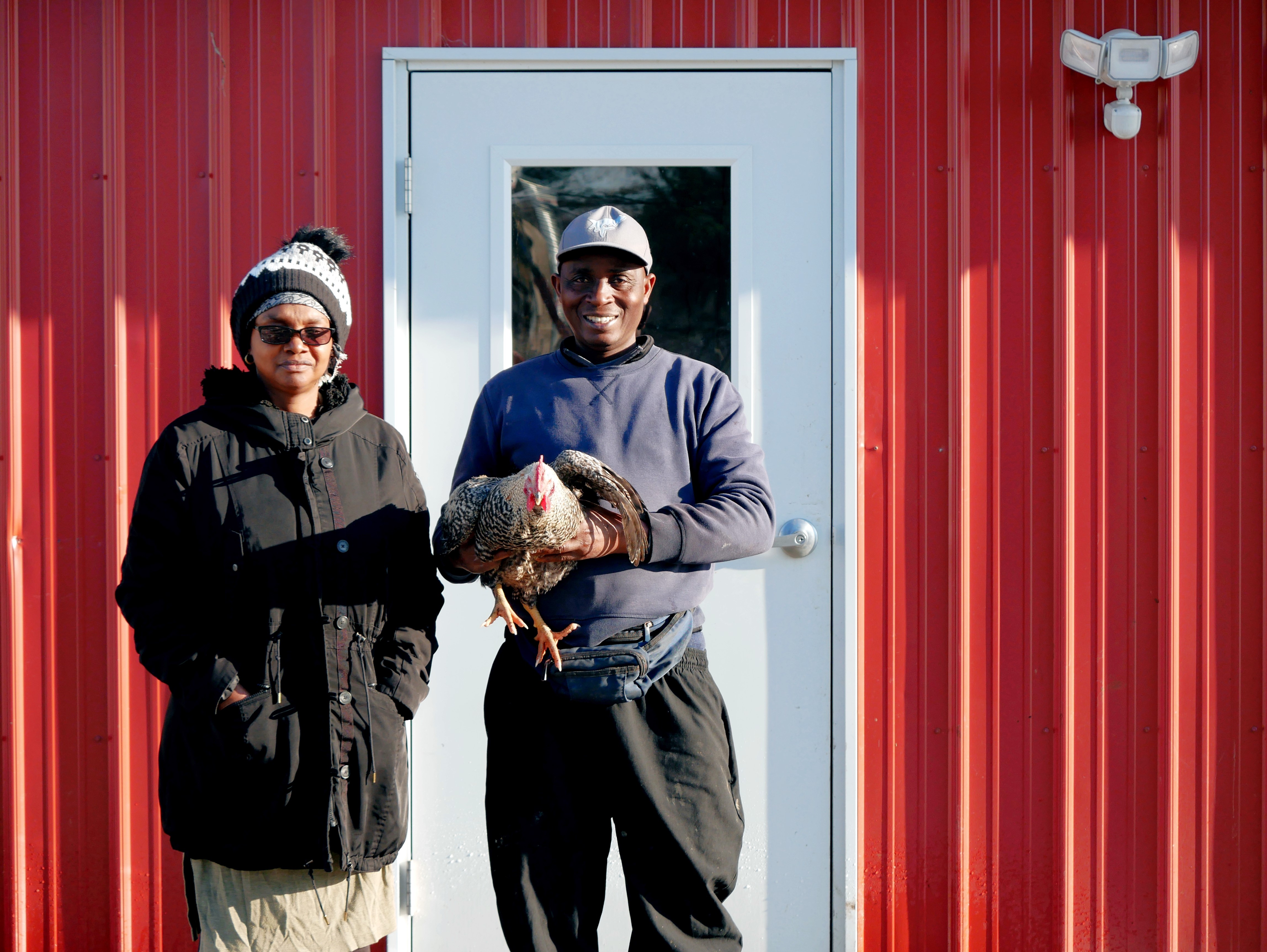 Theogene is holding a chicken and stands with Hyacinthe in front of red building