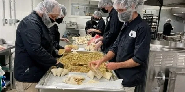 Kitchen staff stand around an industrial table in a line and fill samosas