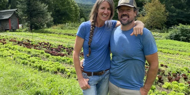 Two people stand together in a hillside farm field with trees and mountains in the background