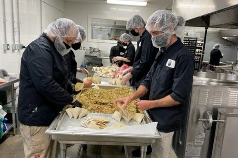 Kitchen staff stand around an industrial table in a line and fill samosas