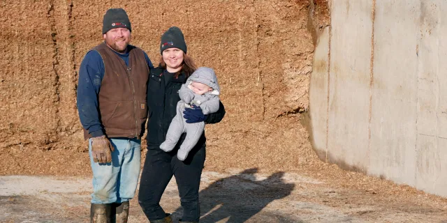 Elijah and Mary White stand in front of feed bunk silo. Mary holds a baby. It is winter.