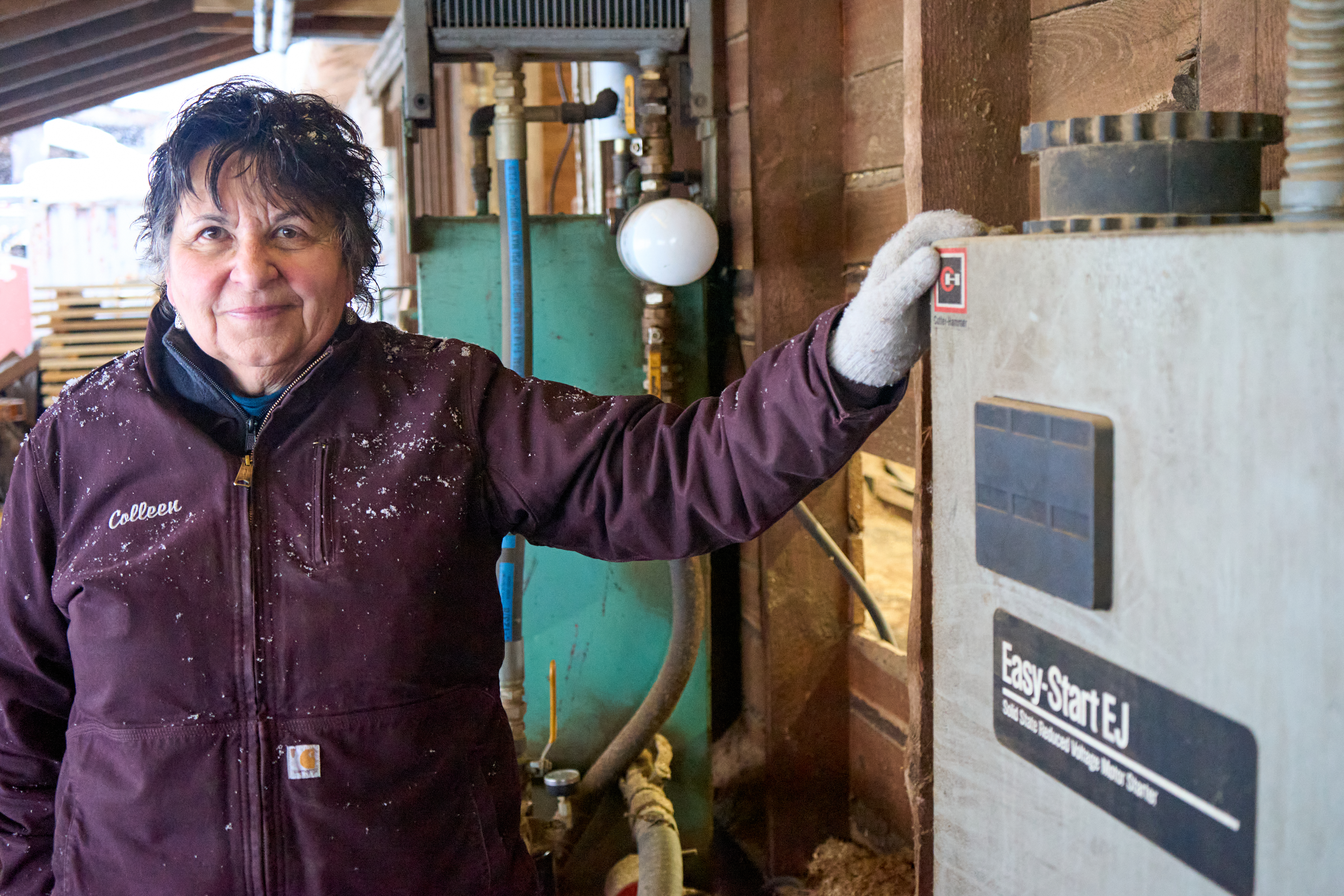 Colleen Goodridge wears a purple coat and stands inside a sawmill next to an electric meter