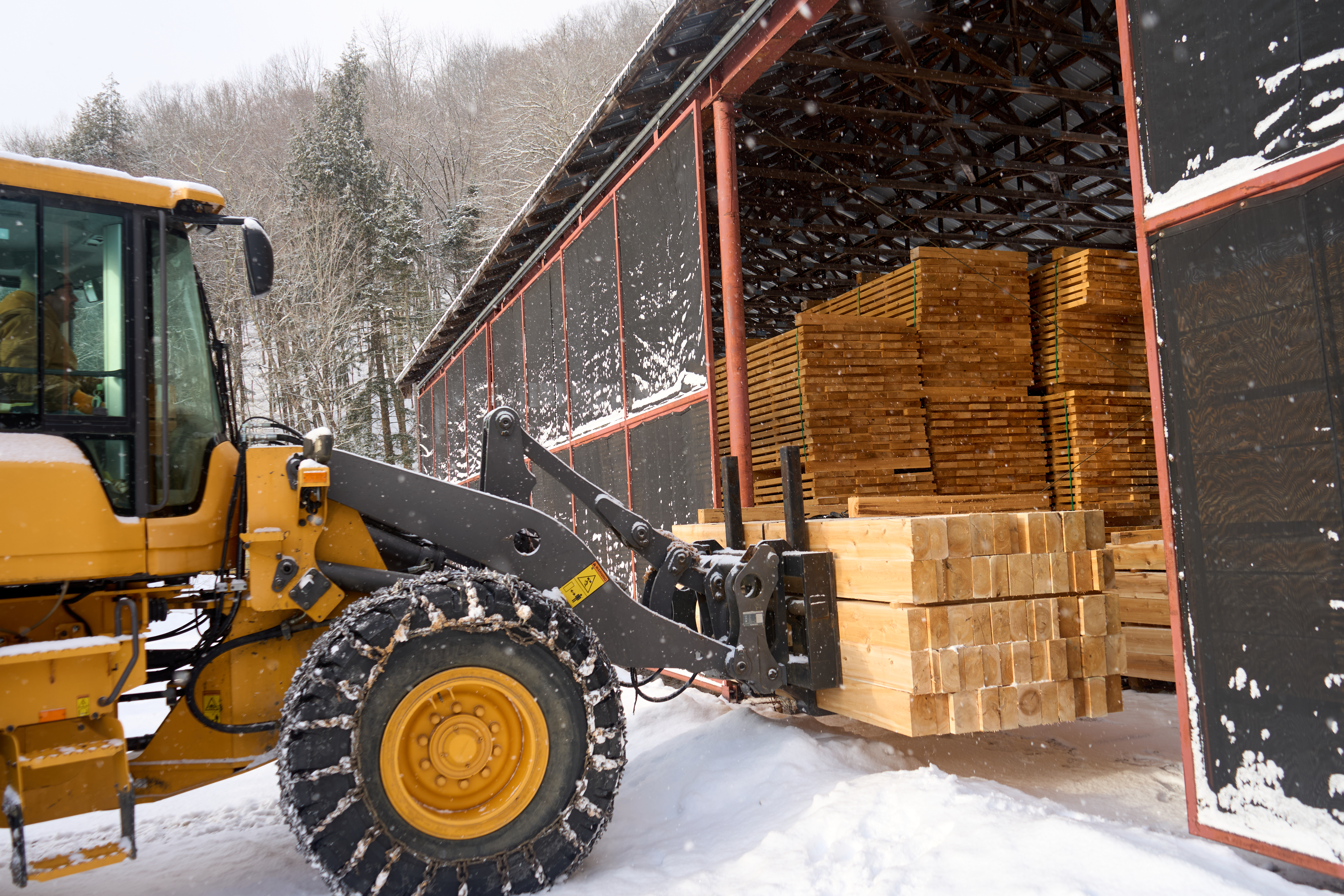 A forklift in the snow moves sawed timbers into a storage area of sawed lumber