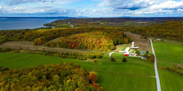 Drone photo of Carman Brook Farm on a partly sunny fall day. There are red farm buildings and silos. There is a lake on the left side of the photo.