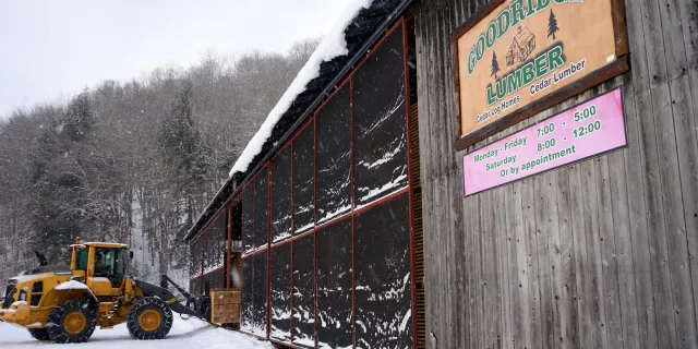 Exterior of sawmill timber storage area in winter. A forklift loads timbers inside. A sign says "Goodridge Lumber" and lists the hours of operation.