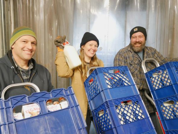 3 people stand with dollies stacked with blue crates with milk bottles. The person in the middle holds up a bottle. People are wearing winter jackets and hats.