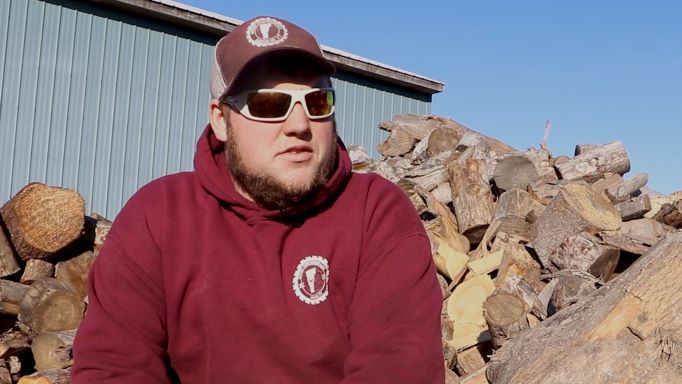 Ethan Gevry wearing a red hoodie, hat, and sunglasses, sitting in front of a pile of firewood