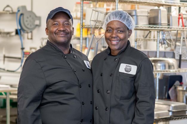Two people (Mel and Damaris Hall) stand next to each other wearing black chef coats that say "Global Village Foods"