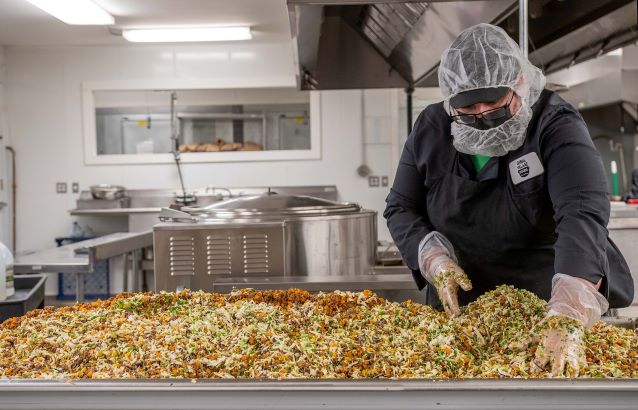 An industrial kitchen counter is covered with samosa filling, and a chef stirs it