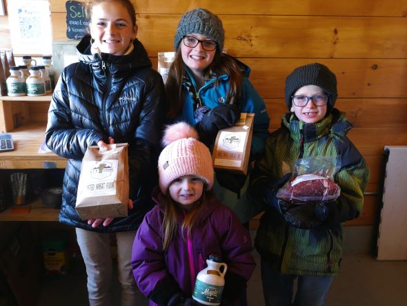 The 4 Gingue children stand in the farm store holding flour, pancake mix, syrup, and meat for sale