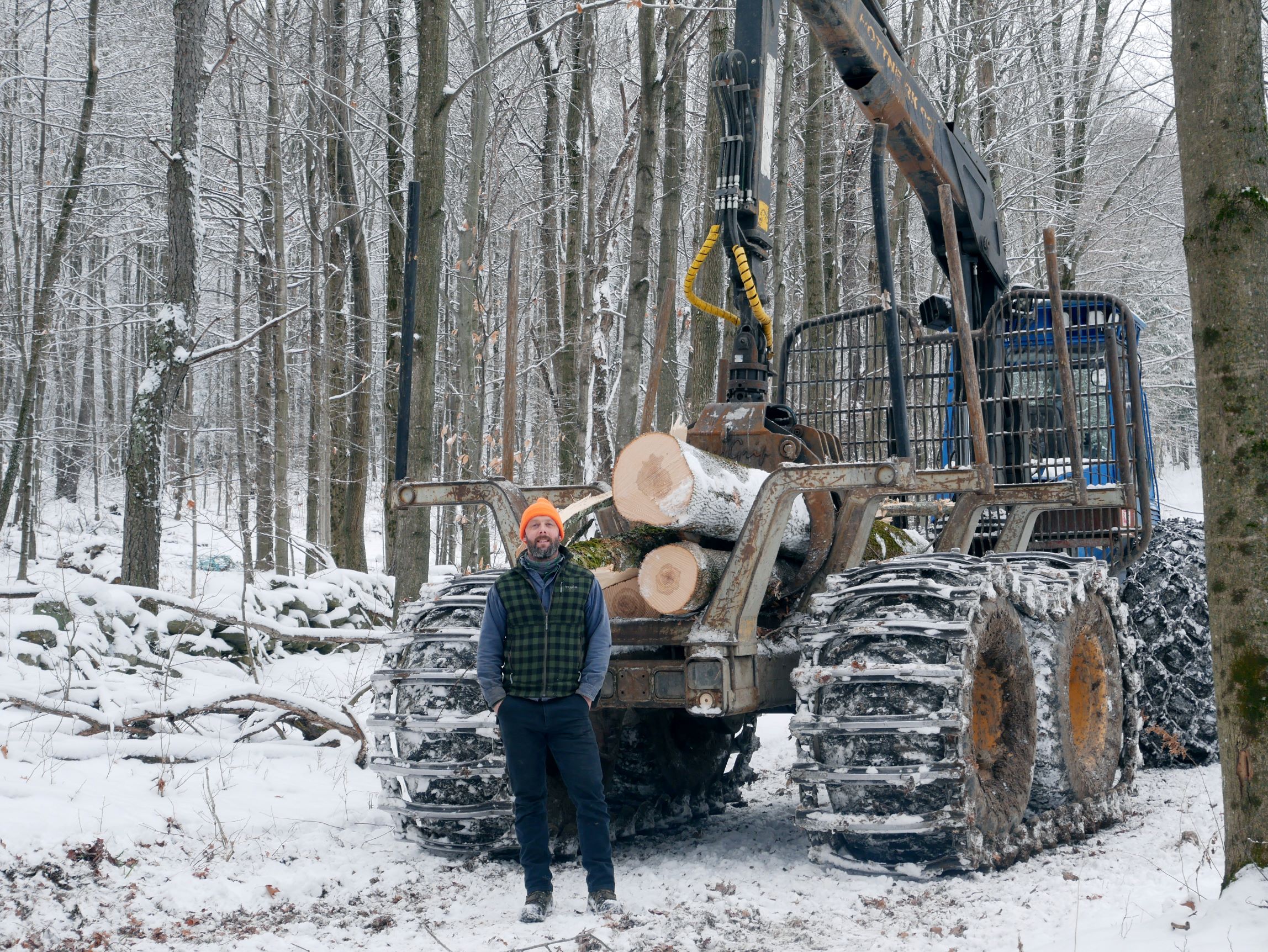 Logging forwarder in the winter with Ben standing in front of it wearing an orange hat