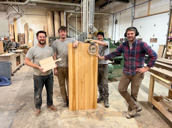 Four men stand in a woodworking shop holding a wood cutting board and butcher block that says "Vermont Butcher Block Company"