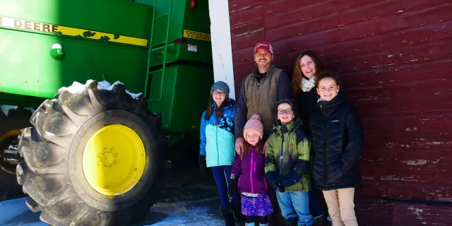 Gingue family of 2 adults and 4 children stand in front of a red barn next to a green tractor