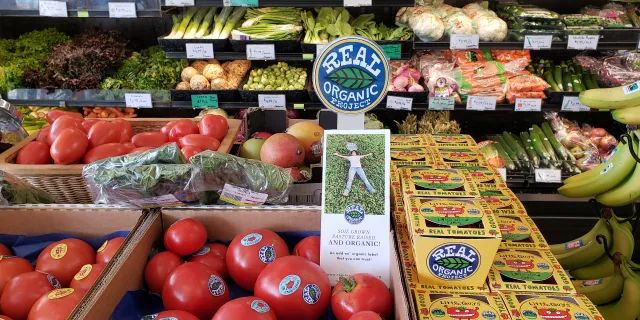 Vegetables on display at a store. There is a sign that says "Real Organic Project" with a green leaf. Some products are also labeled with the sign.